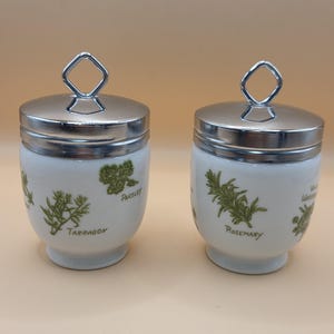May include: Two white ceramic egg coddlers with shiny silver metal lids and diamond-shaped handles. The white bodies feature green botanical illustrations of herbs with the printed text labels Tarragon, Parsley, and Rosemary on the exterior.