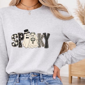 May include: A light gray long-sleeve sweatshirt featuring the word SPOOKY in black and white patterns. The letters O and O are replaced by two ghosts, one wearing a black top hat and bowtie, and the other wearing a white bridal veil and holding a bouquet.