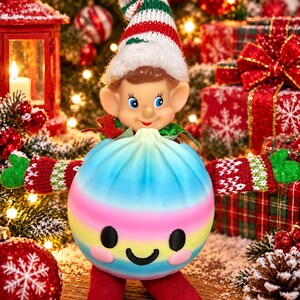 May include: A small elf figurine wearing a knit hat with red, white, and green stripes and a white pom-pom. The elf has pointed ears and holds a round, pastel-coloured dumpling-shaped ornament with a smiling face, pink cheeks, and long red legs.