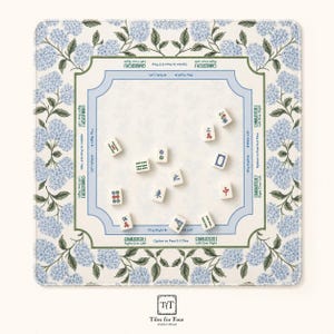 Op de afbeelding: Een vierkante Mahjong-mat met een blauwe hortensia-bloemenrand en een centraal speelveld met gedrukte instructies voor Charleston-zetten. Elf witte Mahjong-tegels met diverse Chinese karakters en symbolen liggen verspreid over de mat.