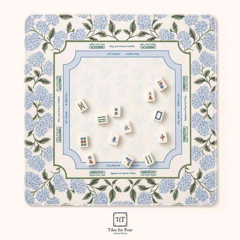 May include: A square Mahjong mat featuring a blue hydrangea floral border and a central playing area with printed instructions for Charleston moves. Eleven white Mahjong tiles with various Chinese characters and symbols are scattered across the mat.