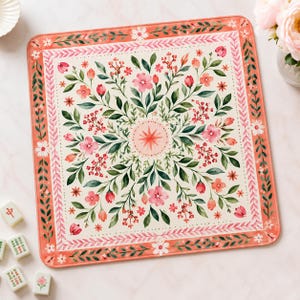 May include: A square mat featuring a cream-colored center with a symmetrical watercolor floral pattern of pink flowers, green leaves, and a central starburst. The border is a coral-orange frame decorated with a repeating leaf and flower motif.