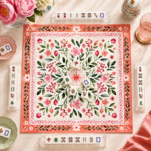 May include: A square mahjong mat featuring a white background with a symmetrical floral pattern in shades of pink, red, and green. The mat is surrounded by four clear tile racks containing mahjong tiles with Chinese characters and symbols.