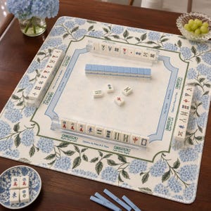 May include: A square mahjong mat featuring a blue and green hydrangea floral pattern on the border. The center includes printed Charleston game instructions and text. Four clear acrylic racks hold white tiles with red, blue, and black symbols.