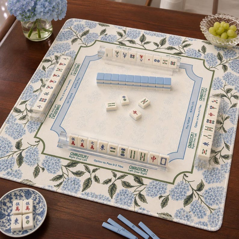 May include: A square mahjong mat featuring a blue and green hydrangea floral pattern on the border. The center includes printed Charleston game instructions and text. Four clear acrylic racks hold white tiles with red, blue, and black symbols.