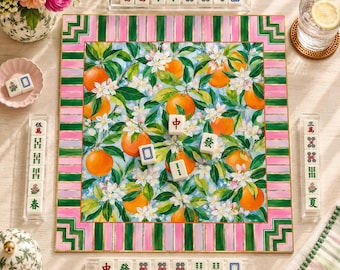May include: A square mahjong board featuring a vibrant orange and white blossom pattern, framed by green and pink stripes. The board includes scattered mahjong tiles with Chinese characters and symbols, alongside three tile racks and a glass of water.