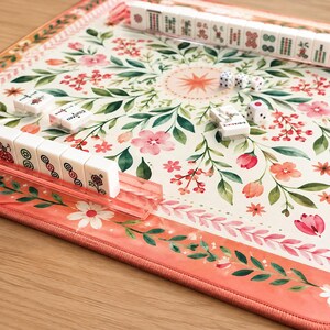May include: A Mahjong game set featuring a rectangular play mat with a cream background, decorated with a floral pattern of pink flowers and green leaves, accompanied by white tiles with red and green symbols and three small white dice.