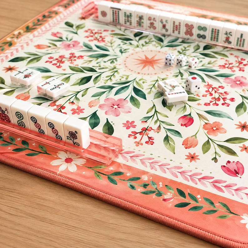 May include: A Mahjong game set featuring a rectangular play mat with a cream background, decorated with a floral pattern of pink flowers and green leaves, accompanied by white tiles with red and green symbols and three small white dice.