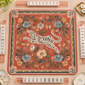 May include: A square mahjong mat featuring a central leaping tiger surrounded by blue and yellow floral patterns on a terracotta background. The mat is bordered by a scalloped green edge and is surrounded by four acrylic tile racks and tiles.