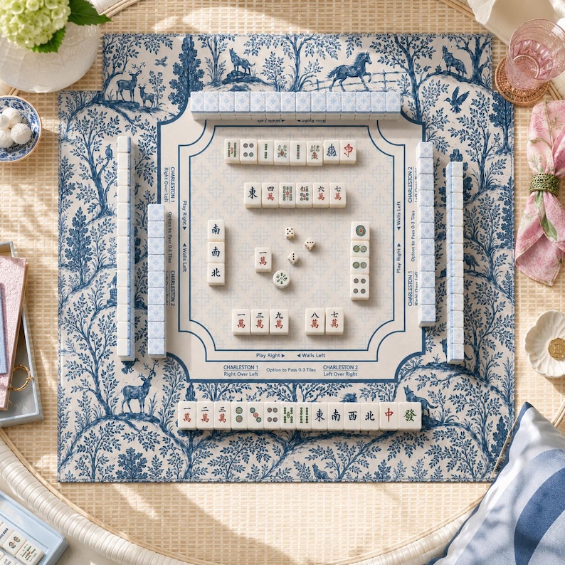 May include: A Mahjong game set featuring a cream-colored play mat with a blue toile de Jouy pattern of trees and animals. The mat includes printed instructions for Charleston moves. Rectangular white tiles with red and black characters are arranged.