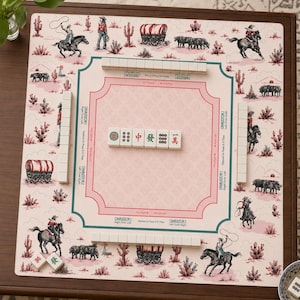 May include: A Western-themed Mahjong set with a pink and white game board featuring cowboys, cacti, and covered wagons. The set includes white tiles with Chinese characters and symbols, and tile racks. The board is set on a brown wooden table.