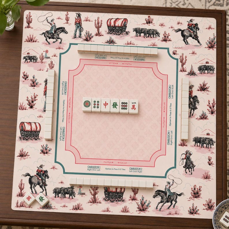 May include: A Western-themed Mahjong set with a pink and white game board featuring cowboys, cacti, and covered wagons. The set includes white tiles with Chinese characters and symbols, and tile racks. The board is set on a brown wooden table.