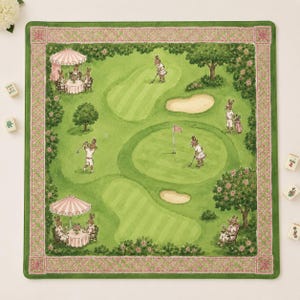 May include: A square fabric mat featuring a green golf course illustration with anthropomorphic rabbits playing golf, lounging under striped umbrellas, and sitting by trees. The mat has a decorative pink and green lattice border pattern.