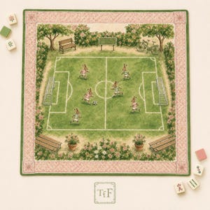 May include: A square fabric mat featuring a printed illustration of rabbits playing soccer on a green field. The scene includes benches, trees, and a bicycle, all framed by a pink and green border. The text TfF is printed at the bottom center.
