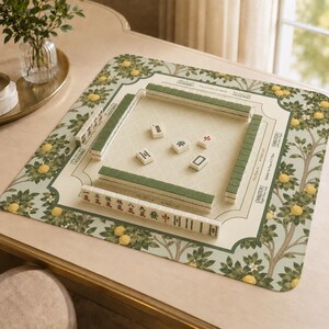 May include: A Mahjong game set on a cream-colored table. The set includes green and white tiles arranged on a patterned mat with a lemon tree design. The mat has a cream-colored center with the words "Charleston" and "Right Door Left" printed on it.