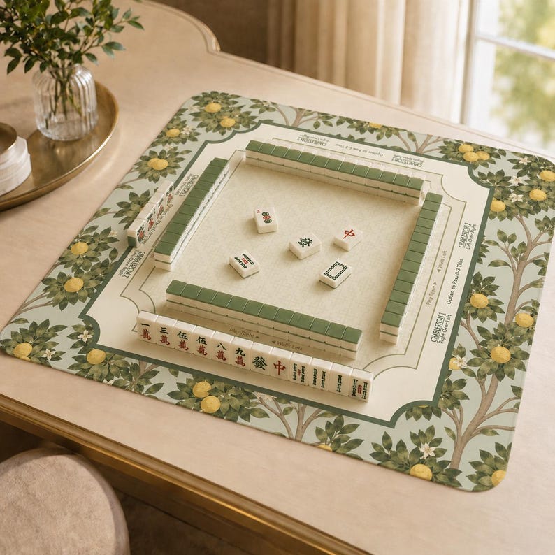 May include: A Mahjong game set on a cream-colored table. The set includes green and white tiles arranged on a patterned mat with a lemon tree design. The mat has a cream-colored center with the words "Charleston" and "Right Door Left" printed on it.