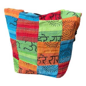 Peut inclure: Un sac à bandoulière en patchwork multicolore composé de carrés de tissu rouges, orange, jaunes, verts et bleus. Le sac est orné de symboles noirs imprimés, incluant le signe Om et des inscriptions en écriture devanagari à motifs répétés.