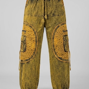 Op de afbeelding: Mosterdgele cargobroek met een donkere acid-wash afwerking, voorzien van een elastische tailleband met trekkoord en grote ronde zijzakken. De zakken zijn versierd met een zwart geometrisch patroon en bevatten kleinere vakken met rits.