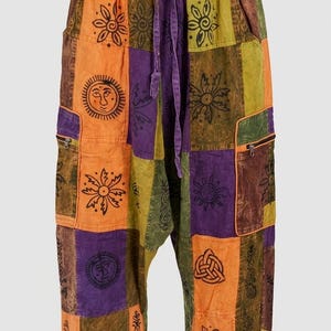 Puede incluir: Pantalones de patchwork con una paleta de colores vibrantes en naranja, morado y verde oliva. La prenda cuenta con una cintura elástica con cordón morado, dos bolsillos laterales con cremalleras y motivos negros estampados de soles y flores.