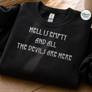 May include: A black crewneck sweatshirt featuring white embroidered text that reads HELL IS EMPTY AND ALL THE DEVILS ARE HERE. The garment has long sleeves, a ribbed neckline, and a white interior brand label at the collar.
