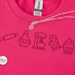 May include: A bright pink sweatshirt featuring dark pink embroidered line art icons of a rolling pin, an apron, a stand mixer, an oven mitt, and a cupcake. The garment has a Gildan brand tag at the neck and a small circular Clover Studios logo.