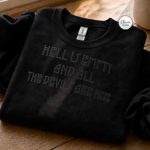 May include: A black crewneck sweatshirt featuring dark, tonal embroidery on the chest that reads, "HELL IS EMPTY AND ALL THE DEVILS ARE HERE." The garment includes a white interior neck label and a small circular logo sticker in the top right corner.