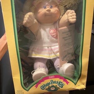 May include: A 1985 Cabbage Patch Kids doll with blonde yarn hair and clear plastic glasses, wearing a white dress with a pink heart patch. The doll is inside its original yellow cardboard box, which includes a birth certificate and adoption papers.