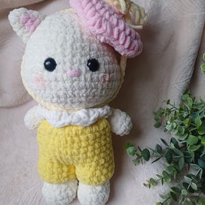 Pode incluir: Um peluche feito &agrave; m&atilde;o em croch&eacute; representando um gato branco com olhos pretos, um pequeno nariz rosa e bochechas rosadas. O gato usa uma boina rosa texturizada com um la&ccedil;o amarelo, um colarinho branco com folhos e jardineiras amarelas.