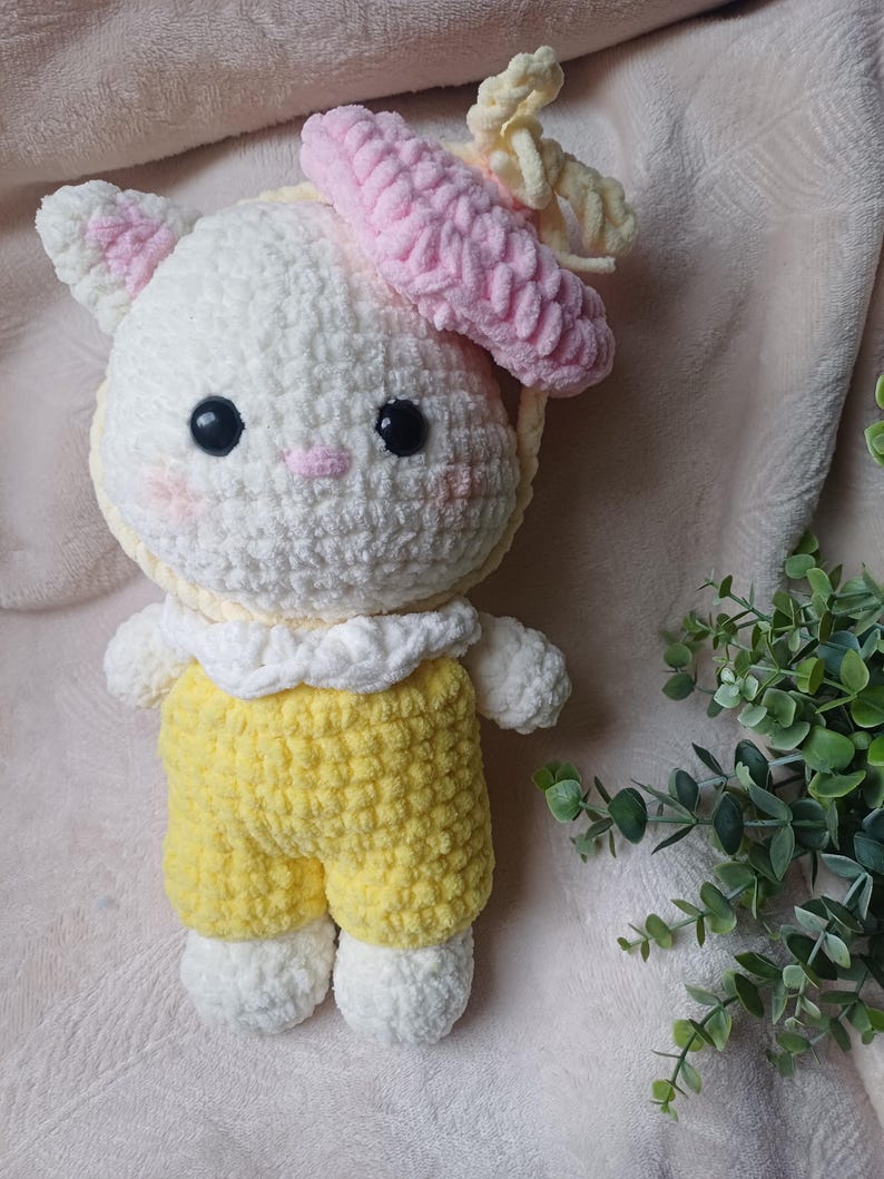 Pode incluir: Um peluche feito &agrave; m&atilde;o em croch&eacute; representando um gato branco com olhos pretos, um pequeno nariz rosa e bochechas rosadas. O gato usa uma boina rosa texturizada com um la&ccedil;o amarelo, um colarinho branco com folhos e jardineiras amarelas.