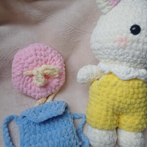 Pode incluir: Um animal de pel&uacute;cia em croch&ecirc; branco com orelhas e bochechas cor-de-rosa, vestindo um macac&atilde;o amarelo. Ao lado, h&aacute; uma pequena mochila de croch&ecirc; azul-claro com aba e um acess&oacute;rio redondo de croch&ecirc; rosa com um la&ccedil;o cor de creme.