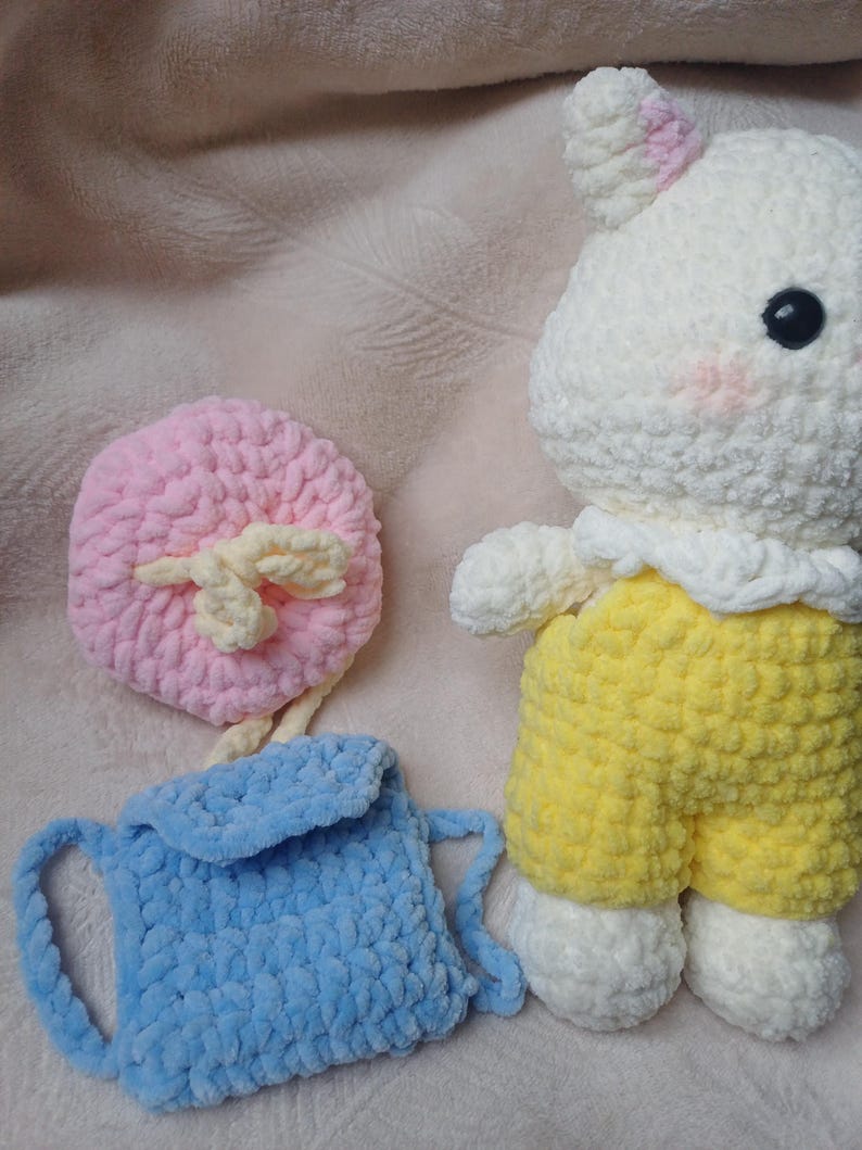 Pode incluir: Um animal de pel&uacute;cia em croch&ecirc; branco com orelhas e bochechas cor-de-rosa, vestindo um macac&atilde;o amarelo. Ao lado, h&aacute; uma pequena mochila de croch&ecirc; azul-claro com aba e um acess&oacute;rio redondo de croch&ecirc; rosa com um la&ccedil;o cor de creme.