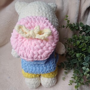 Pode incluir: Um brinquedo de pel&uacute;cia de croch&ecirc; branco vestindo uma camisa azul e cal&ccedil;as amarelas. O brinquedo tem uma mochila redonda de croch&ecirc; rosa com um la&ccedil;o cor de creme preso nas costas. A pel&uacute;cia &eacute; feita de fio macio e texturizado com cabe&ccedil;a arredondada.
