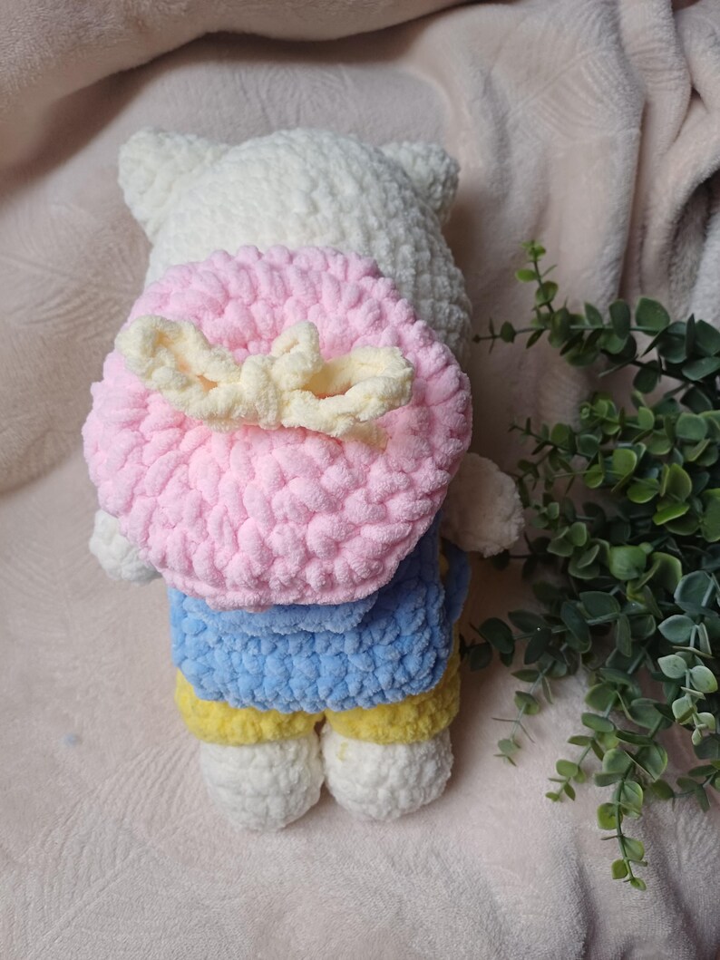 Pode incluir: Um brinquedo de pel&uacute;cia de croch&ecirc; branco vestindo uma camisa azul e cal&ccedil;as amarelas. O brinquedo tem uma mochila redonda de croch&ecirc; rosa com um la&ccedil;o cor de creme preso nas costas. A pel&uacute;cia &eacute; feita de fio macio e texturizado com cabe&ccedil;a arredondada.