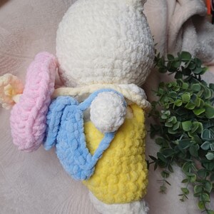 Pode incluir: Um peluche branco feito em croch&eacute; com um acabamento de fio texturizado. A figura usa jardineiras amarelas e transporta uma mochila azul-clara. Um acess&oacute;rio circular em croch&eacute; cor-de-rosa est&aacute; preso na lateral. O peluche tem uma cabe&ccedil;a redonda.