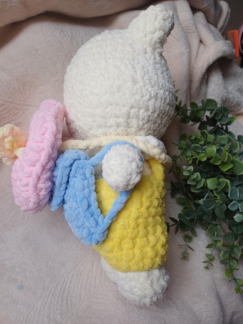 Pode incluir: Um peluche branco feito em croch&eacute; com um acabamento de fio texturizado. A figura usa jardineiras amarelas e transporta uma mochila azul-clara. Um acess&oacute;rio circular em croch&eacute; cor-de-rosa est&aacute; preso na lateral. O peluche tem uma cabe&ccedil;a redonda.
