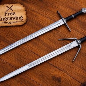May include: Two steel swords with black wrapped handles and metal crossguards. One sword features a circular pommel, while the other has a unique T-shaped pommel. A parchment-style sign nearby displays the text Free Engraving with sword icons.