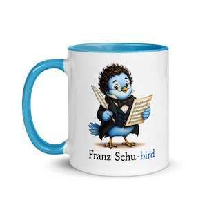 May include: A white ceramic mug with a bright blue handle and interior rim. The mug features a cartoon illustration of a blue bird dressed in a black tuxedo, bow tie, and glasses, holding a quill and sheet music. The text reads Franz Schu-bird.