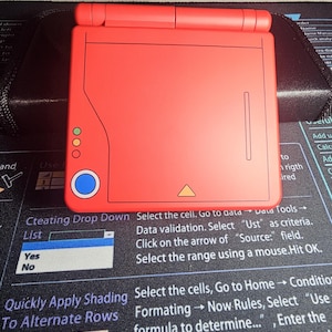 May include: A red handheld gaming device with a flip-up screen. The device has a square design with a button layout on the left side, including a blue circle and three smaller circles. A yellow triangle is also visible.