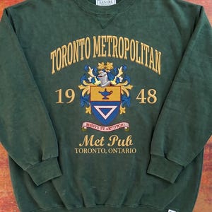 May include: A dark green crewneck sweatshirt featuring the embroidered text Toronto Metropolitan, the numbers 19 and 48, and a crest with a lamp and triangle. Below the crest is a banner with Mente et Artificio, Met Pub, and Toronto, Ontario.