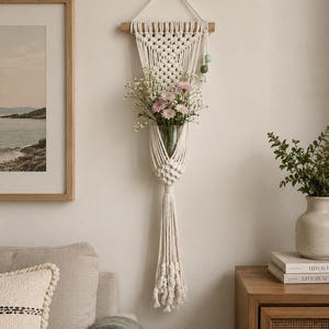 May include: A cream-coloured macrame wall hanging featuring a wooden dowel, intricate knotted patterns, and a central glass vase holding pink and white flowers. The piece includes a long fringe at the bottom and two small green wooden beads.