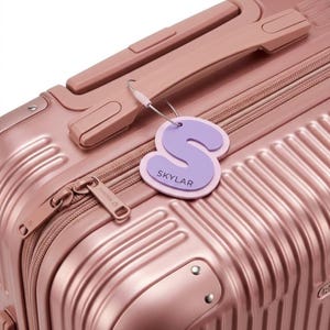 May include: A metallic rose gold suitcase with a ribbed exterior and a top handle. Attached to the handle is a light purple luggage tag shaped like the letter S, featuring the name SKYLAR printed in dark lettering on the lower portion.