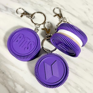 Puede incluir: Tres llaveros con forma de galleta morada y cierres de mosquetón de metal plateado. Dos llaveros presentan el texto BTS en relieve en la parte frontal, mientras que uno muestra un logotipo geométrico. Las galletas tienen un borde estriado.