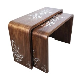 May include: A set of two nesting tables crafted from dark brown wood with a curved waterfall edge design. The surfaces and side panels feature intricate white floral inlay patterns that extend across the top and down the front legs of each table.
