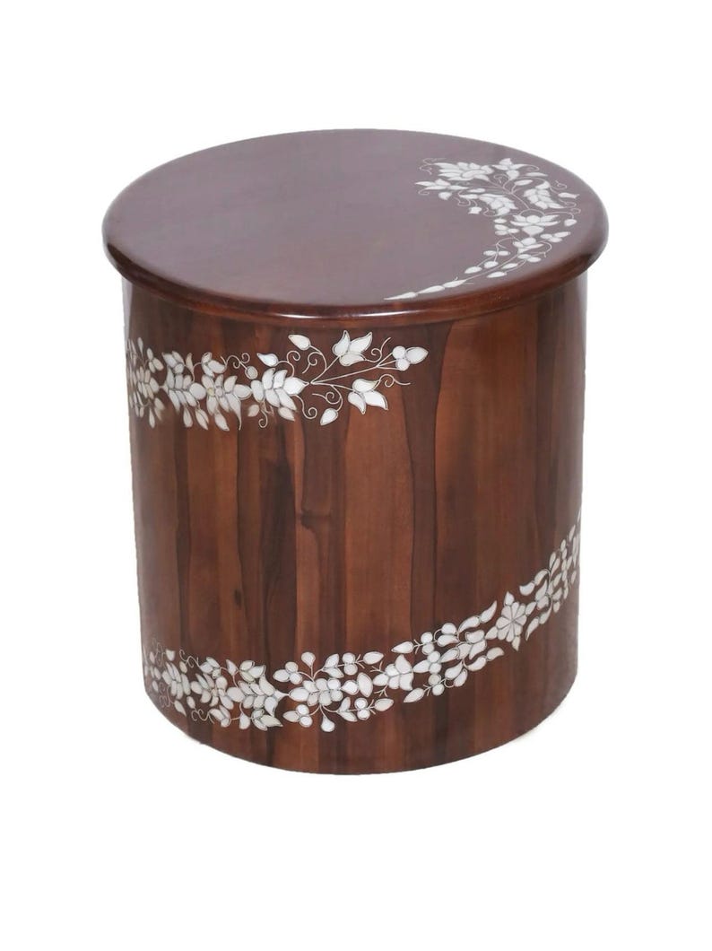 K&ouml;nnte beinhalten: Ein zylindrischer Beistelltisch aus Holz mit einer glatten, dunkelbraunen Oberfl&auml;che. Das M&ouml;belst&uuml;ck ist mit einem eingelegten wei&szlig;en Blumenrankenmuster verziert, das sich um die Seite windet und &uuml;ber den oberen Rand erstreckt.