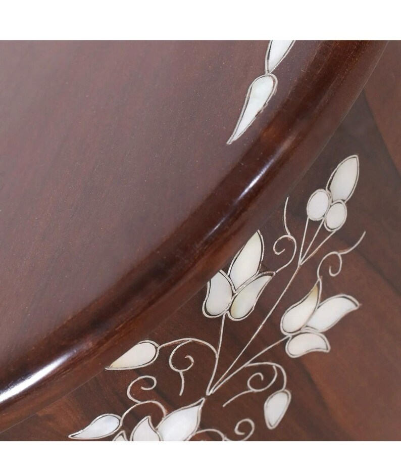 K&ouml;nnte beinhalten: Eine dunkelbraune Holzoberfl&auml;che mit einem eingelegten Blumenmuster aus wei&szlig;em, schillerndem Perlmutt. Das Design besteht aus zarten Stielen, Bl&auml;ttern und Bl&uuml;tenknospen, die in einer flie&szlig;enden, organischen Komposition auf dem Holz angeordnet sind.