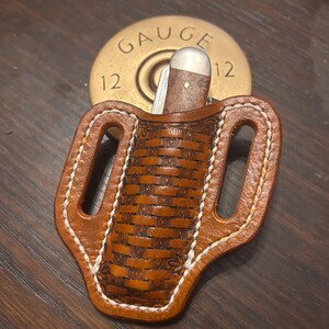 May include: A brown leather knife sheath with a woven design and white stitching. It holds a small folding knife with a wooden handle. A brass 12-gauge shotgun shell casing is behind the sheath.
