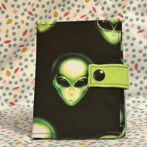 May include: A rectangular fabric wallet featuring a black background with a repeating pattern of green alien heads. The wallet includes a lime green fabric strap closure secured with a single black snap button centered on the right side of the item.