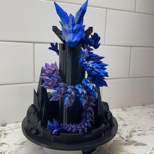 Op de afbeelding: Een 3D-geprinte fantasiedraaksculptuur in blauwe en paarse tinten. De draak staat op een zwarte torenstructuur met een cirkelvormige basis. Het lichaam van de draak is versierd met kristalachtige stekels.