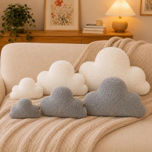 May include: A collection of six cloud-shaped pillows in varying sizes, featuring three white pillows and three gray pillows. Each pillow is crafted from a textured, fuzzy fabric with a soft, rounded silhouette, arranged on a beige knit blanket.