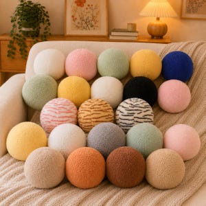 May include: A collection of twenty round, textured, sherpa-style throw pillows arranged on a sofa. The pillows feature a variety of solid colors including white, pink, sage green, yellow, blue, black, gray, orange, and brown, plus two striped patterns.